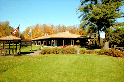 A wooden clubhouse with a large lawn and a small gazebo under a clear blue sky, surrounded by autumn trees.