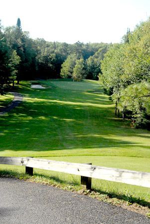 A golf course fairway descends toward a distant green framed by trees, with a wooden barrier in the foreground.