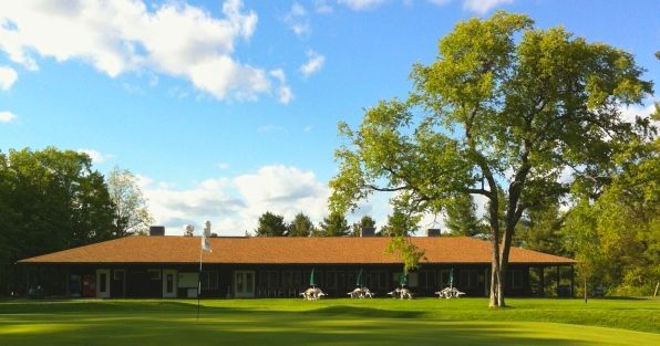 A low, single-story clubhouse with a brown roof sits behind a vibrant green golf course under a bright blue sky.