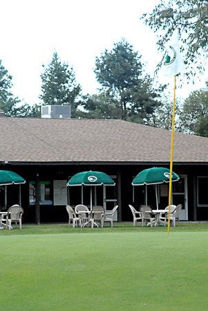 A golf green with a flag leads to a building with outdoor seating under three green umbrellas.