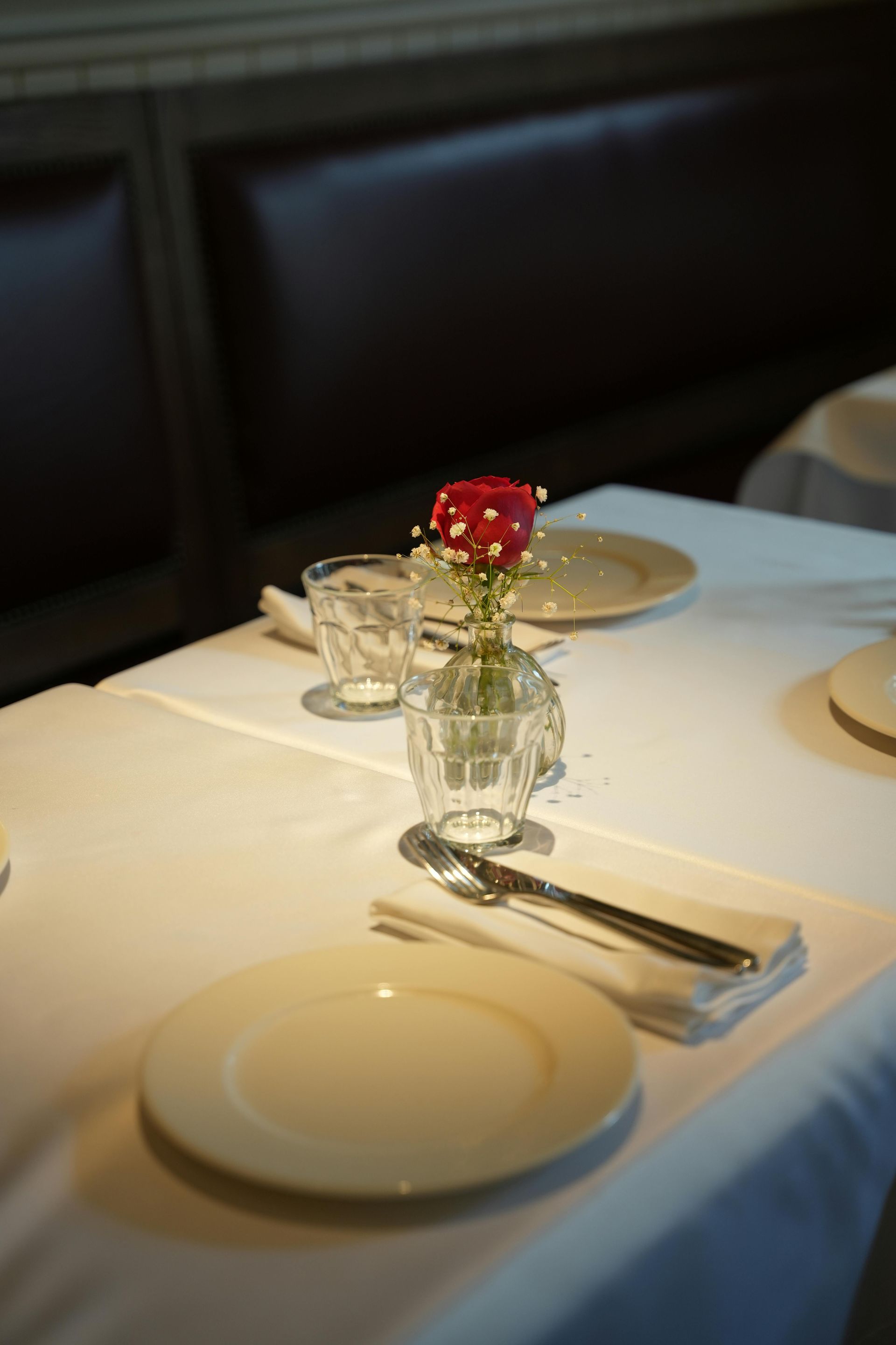 A formal dining table set with a white cloth, plates, silverware, and a small glass vase holding a single red rose.