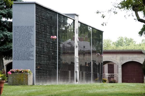 Glass memorial wall with engraved names beside a stone building and flower bed