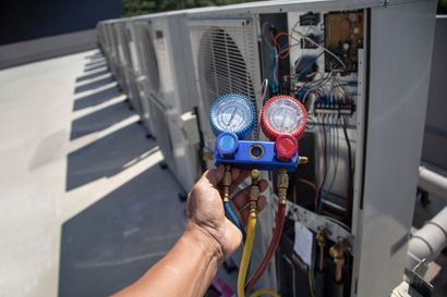 Hand holding HVAC manifold gauge set to an outdoor air conditioning unit with exposed wiring.