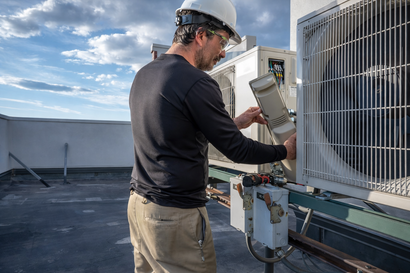 Technician in hard hat inspecting rooftop HVAC unit under a cloudy sky