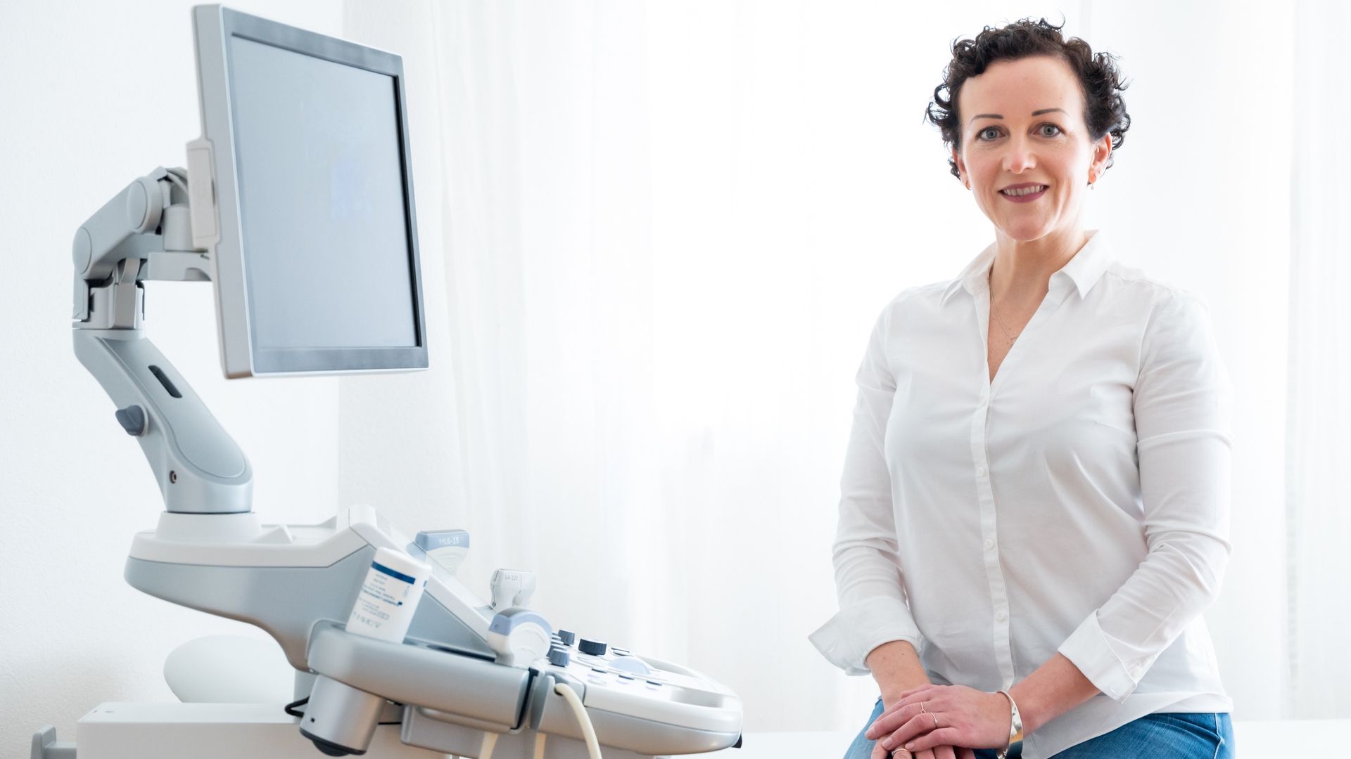 Eine Frau steht neben einem Ultraschallgerät in einer hellen Arztpraxis und lächelt in die Kamera.