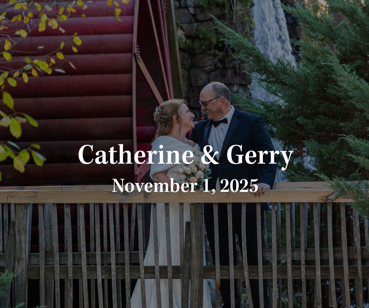 A couple in wedding attire stands on a wooden bridge in front of a water wheel with text reading
