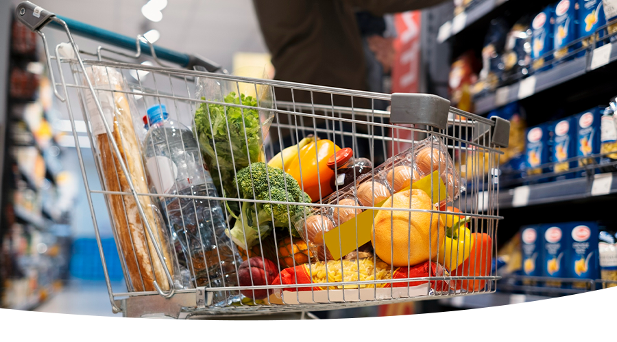 Shopping cart filled with groceries, including bread, vegetables, eggs, and water bottle, in a supermarket aisle.