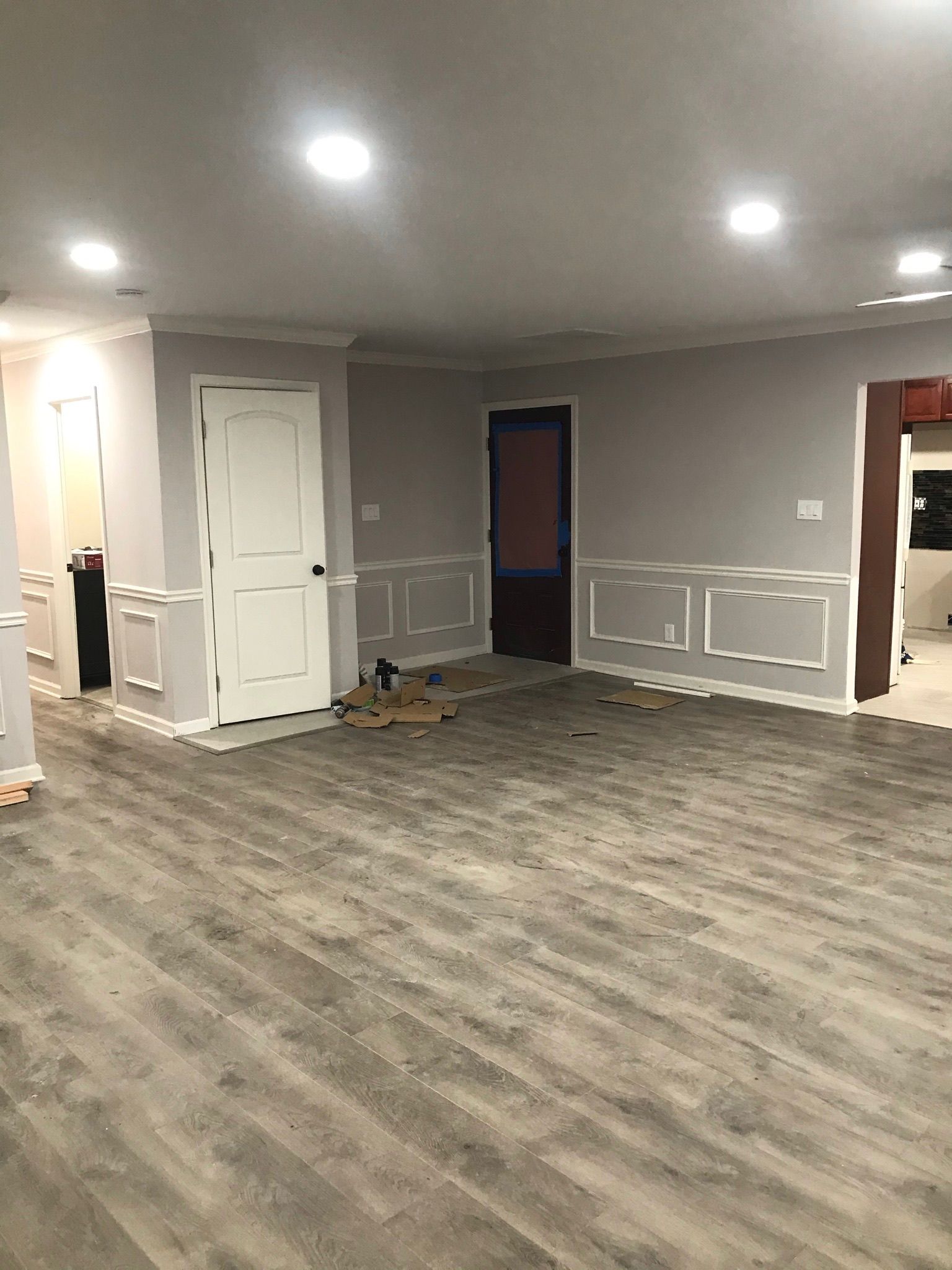 An unfinished room with gray walls, wainscoting, a white door, and a wooden floor, illuminated by recessed lighting.