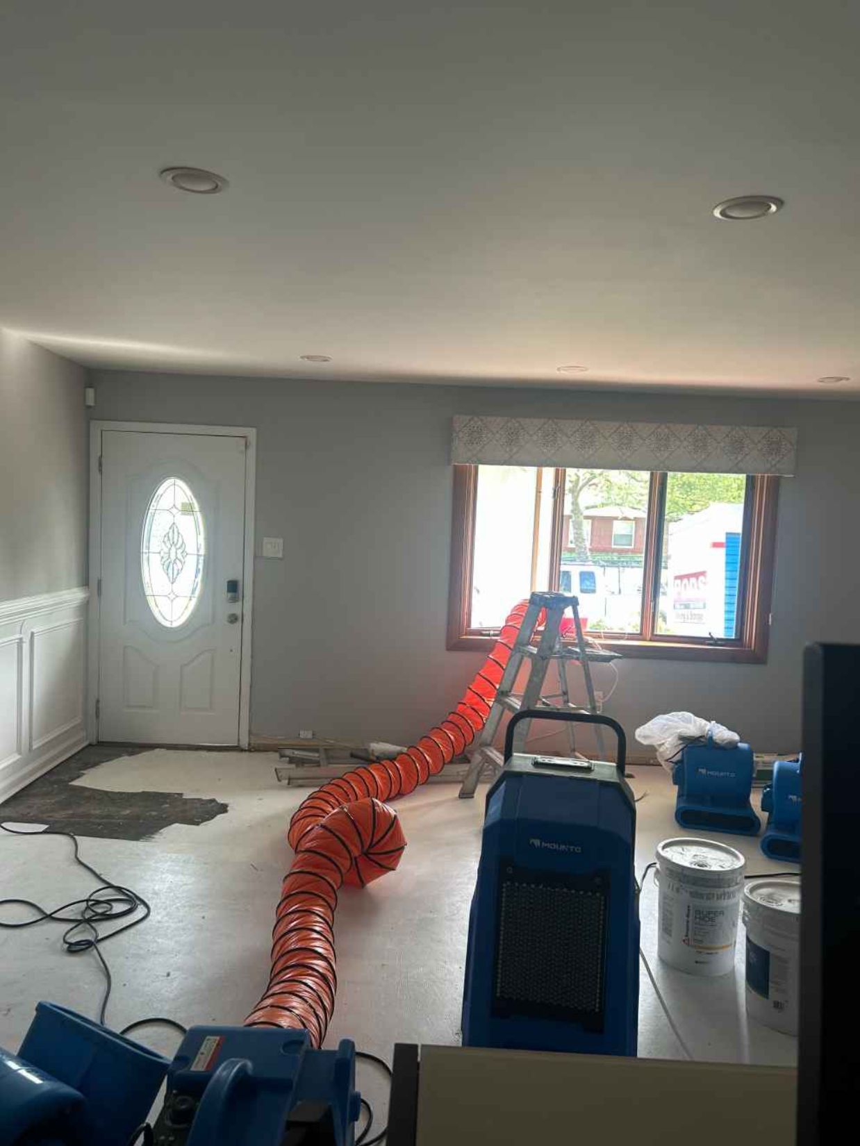 Flood-damaged room with a large orange air duct, dehumidifier, and restoration equipment near the window.