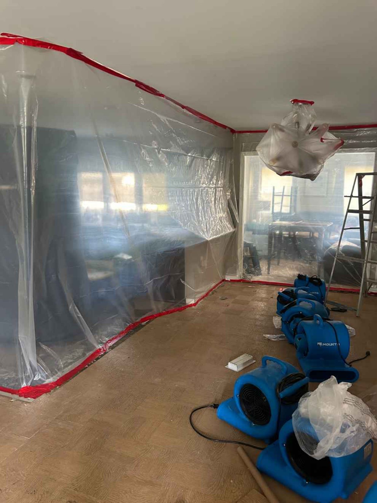 Room sealed with plastic sheeting and red tape; blue fans on the floor during renovation work.