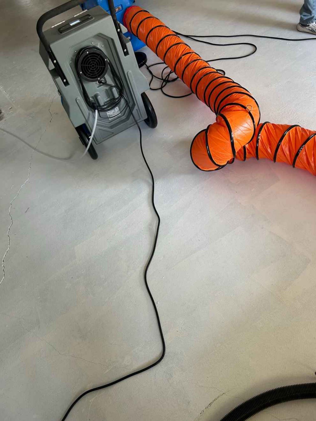 Orange flexible duct attached to a machine on a floor, with a black cable trailing nearby.