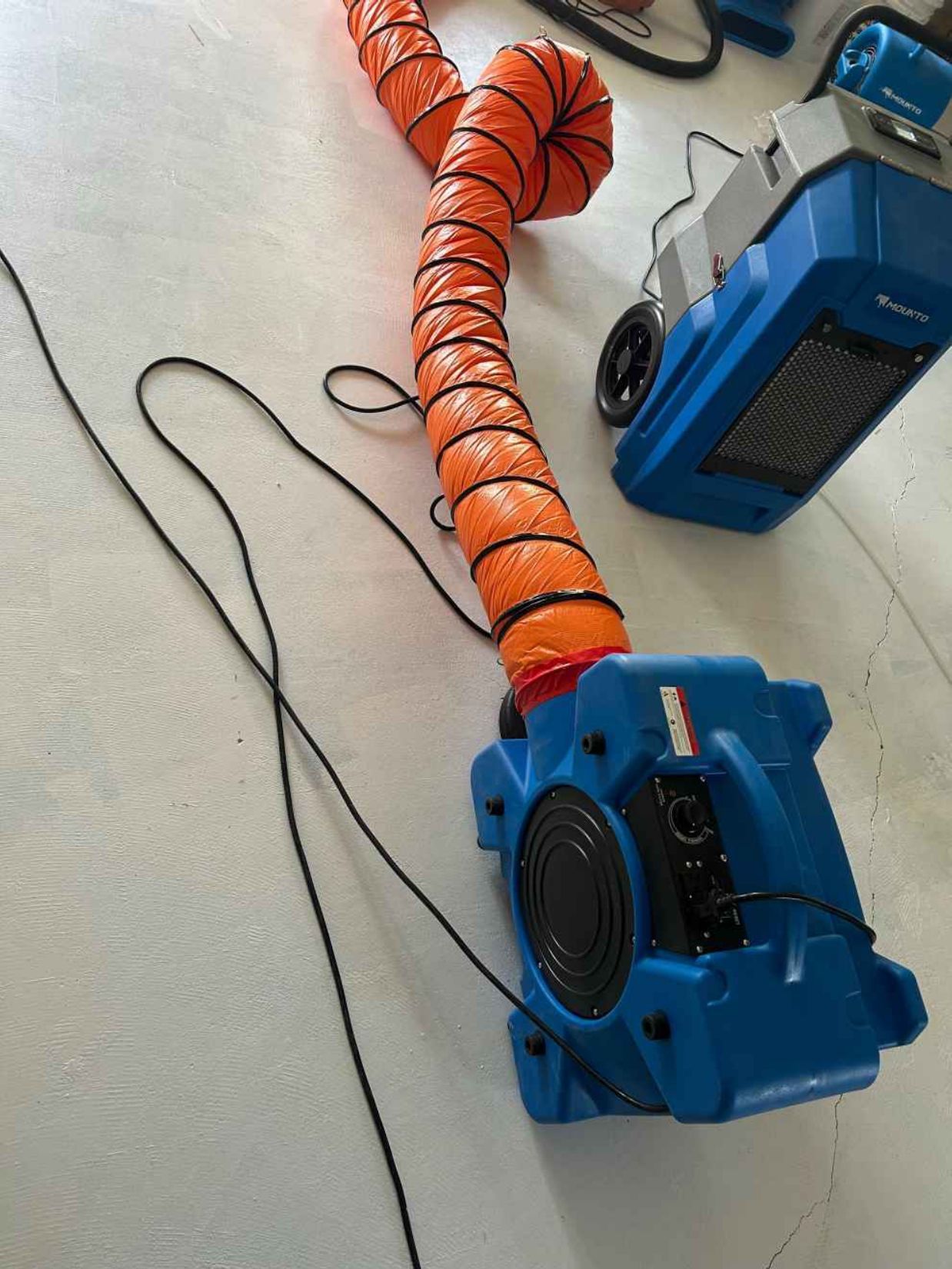 Blue industrial blower attached to an orange ventilation hose on a concrete floor.