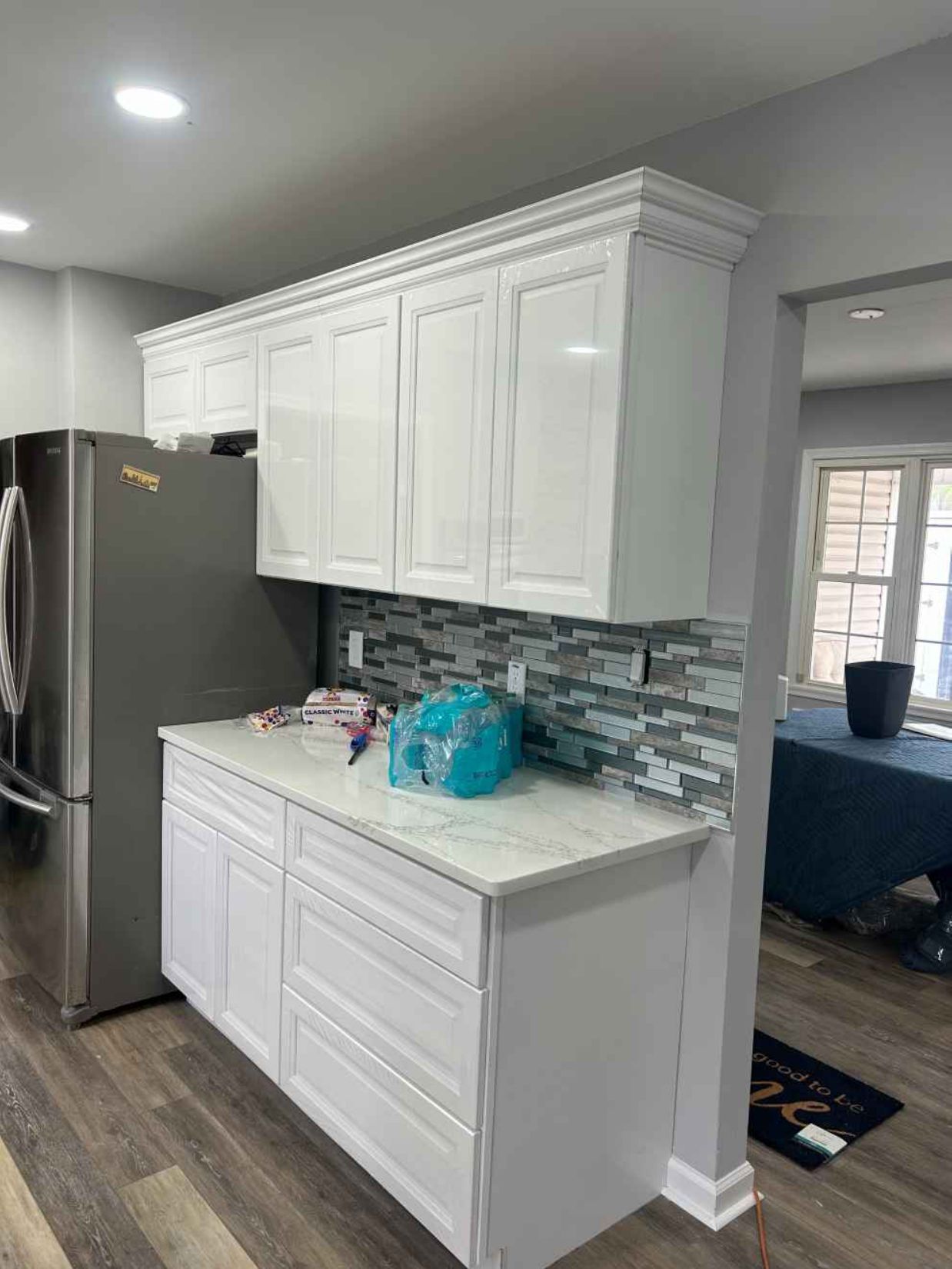 White kitchen cabinets and countertop beside a stainless-steel fridge, with a mosaic tile backsplash.