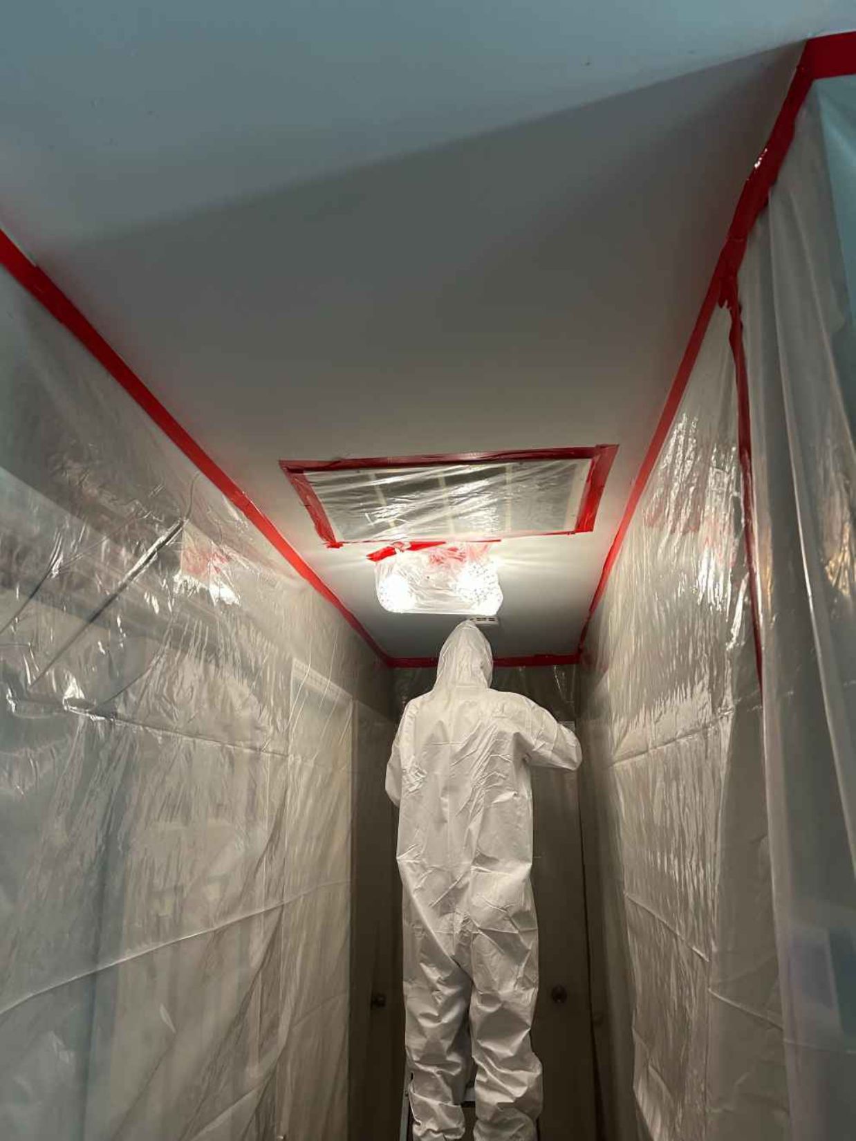 Person in white protective suit inspecting a plastic-covered hallway with red tape and ceiling vent.