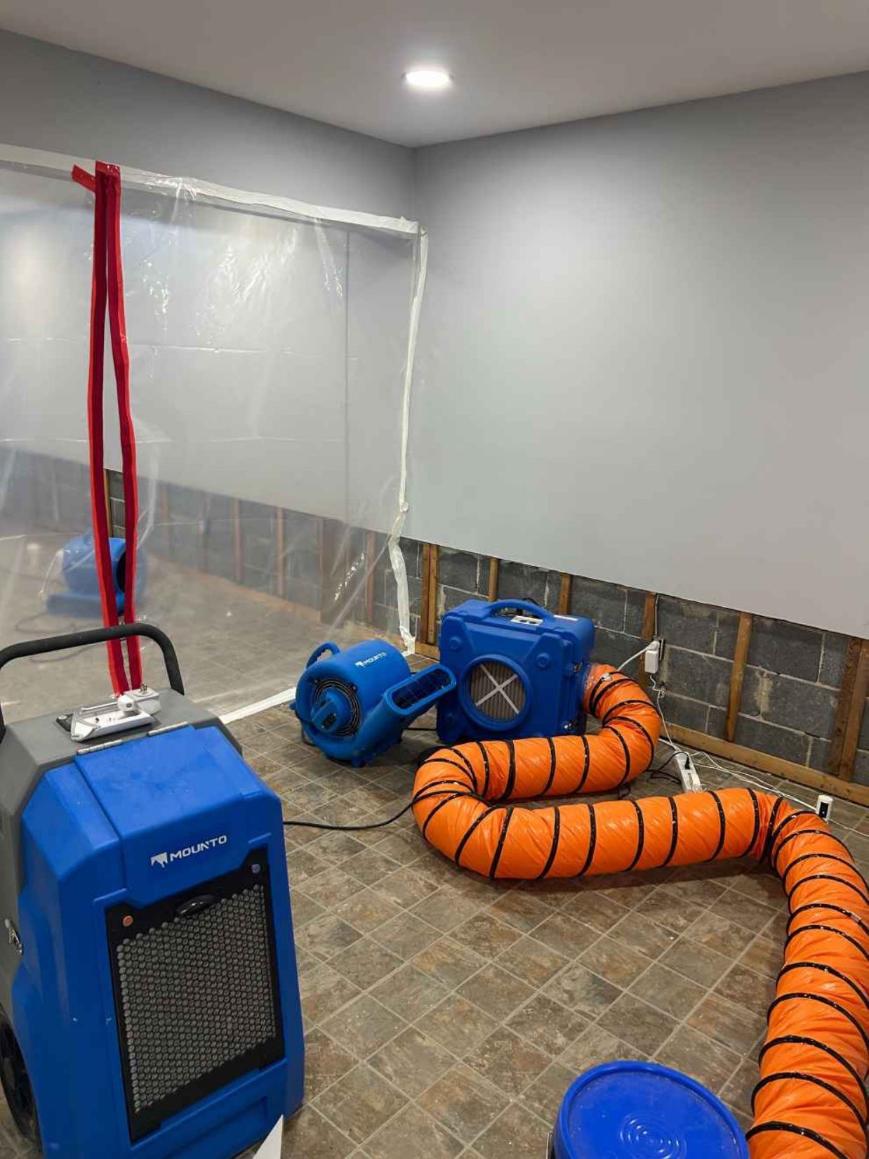 Blue industrial air mover and orange hose in a wet room with a plastic barrier and fan/dryer equipment.
