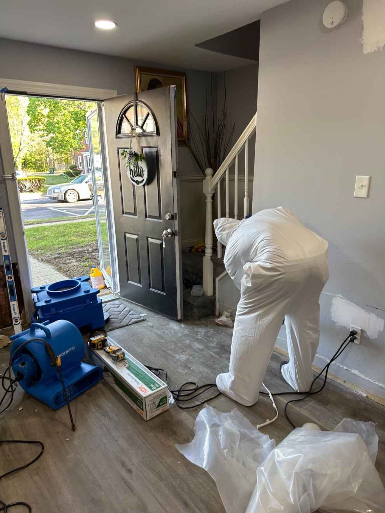 Person in white cleaning equipment near open front door with fan and supplies in a home entryway.