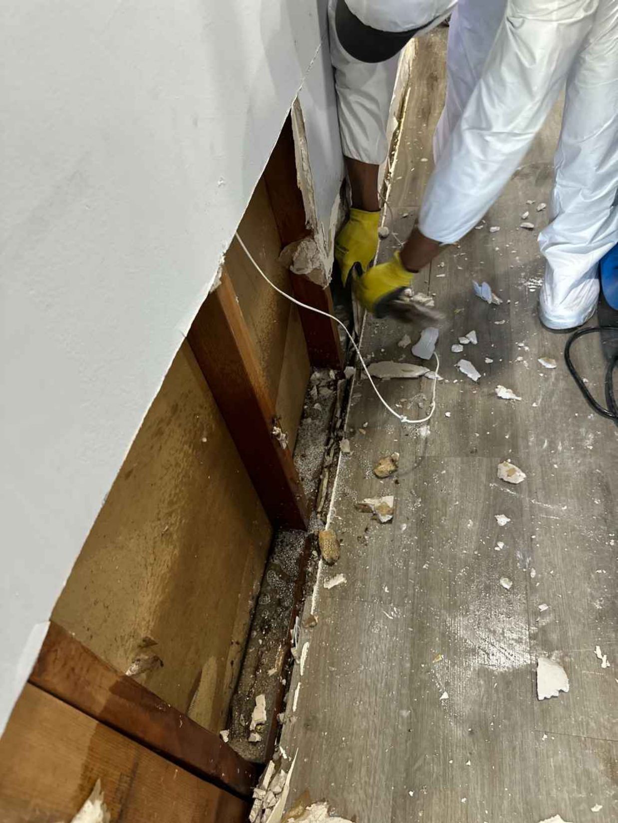 Workers using tools to clear debris along a damaged wall and floor inside a room