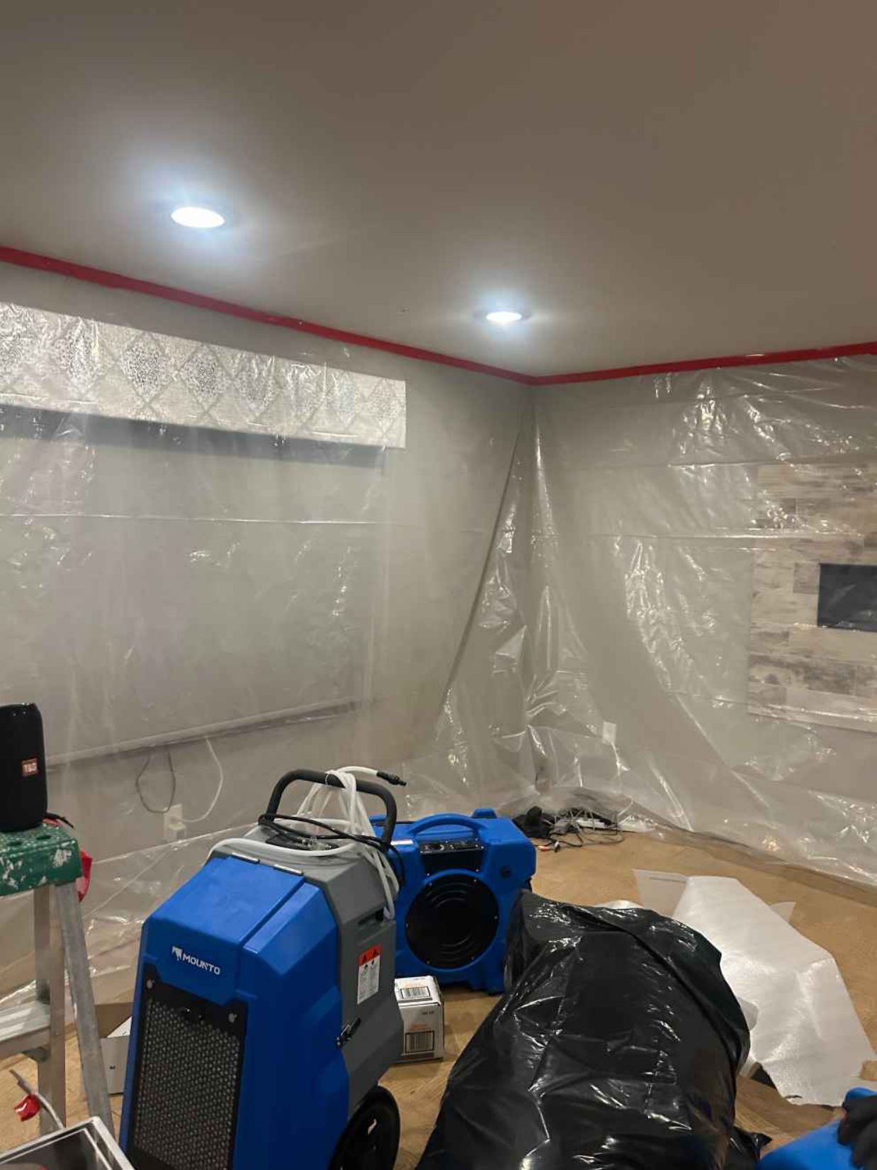 Plastic-covered room with blue construction fans and equipment, suggesting renovation or drying work