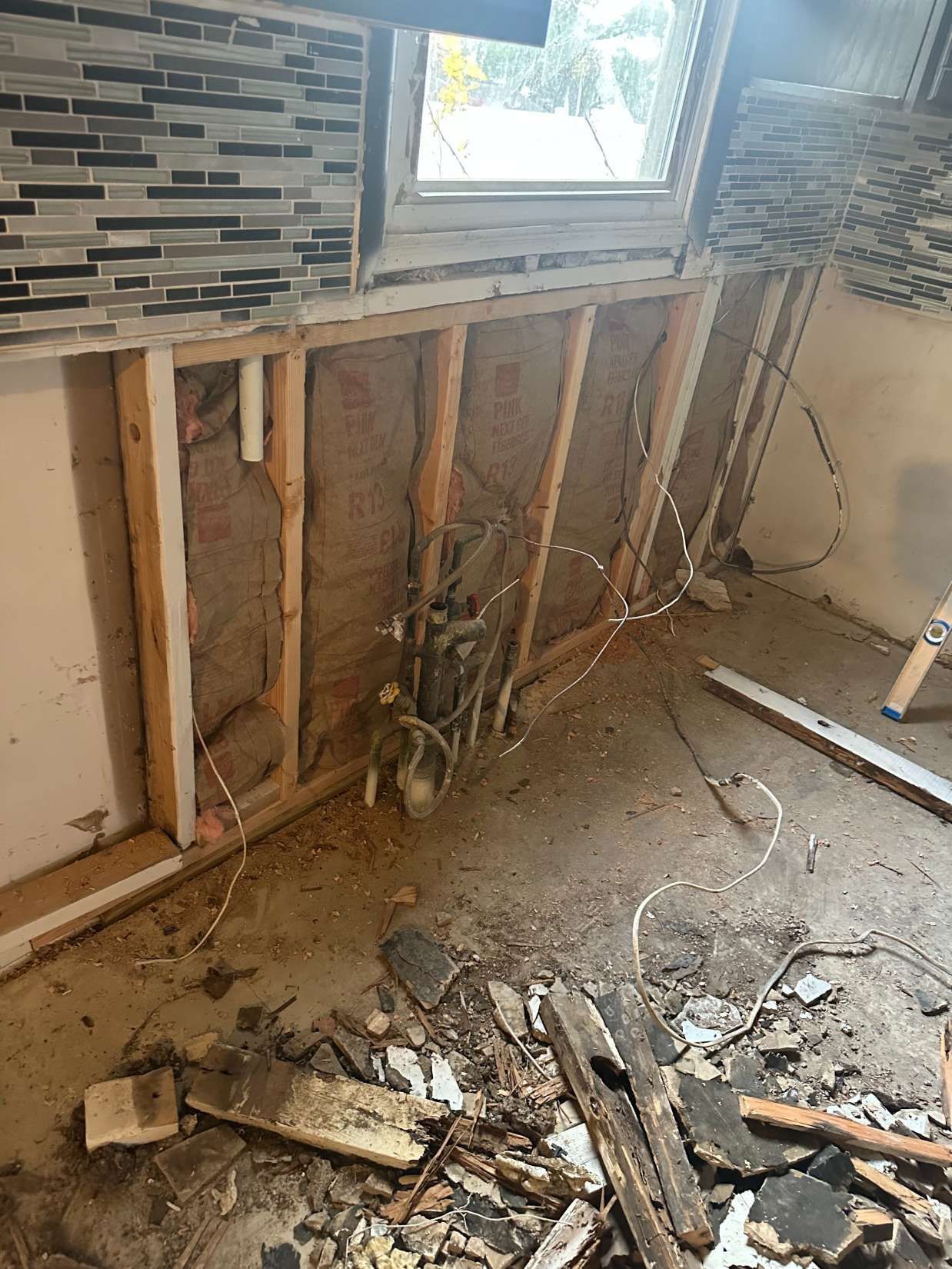Partially demolished room with exposed wall studs, tile backsplash, window, and debris on the floor