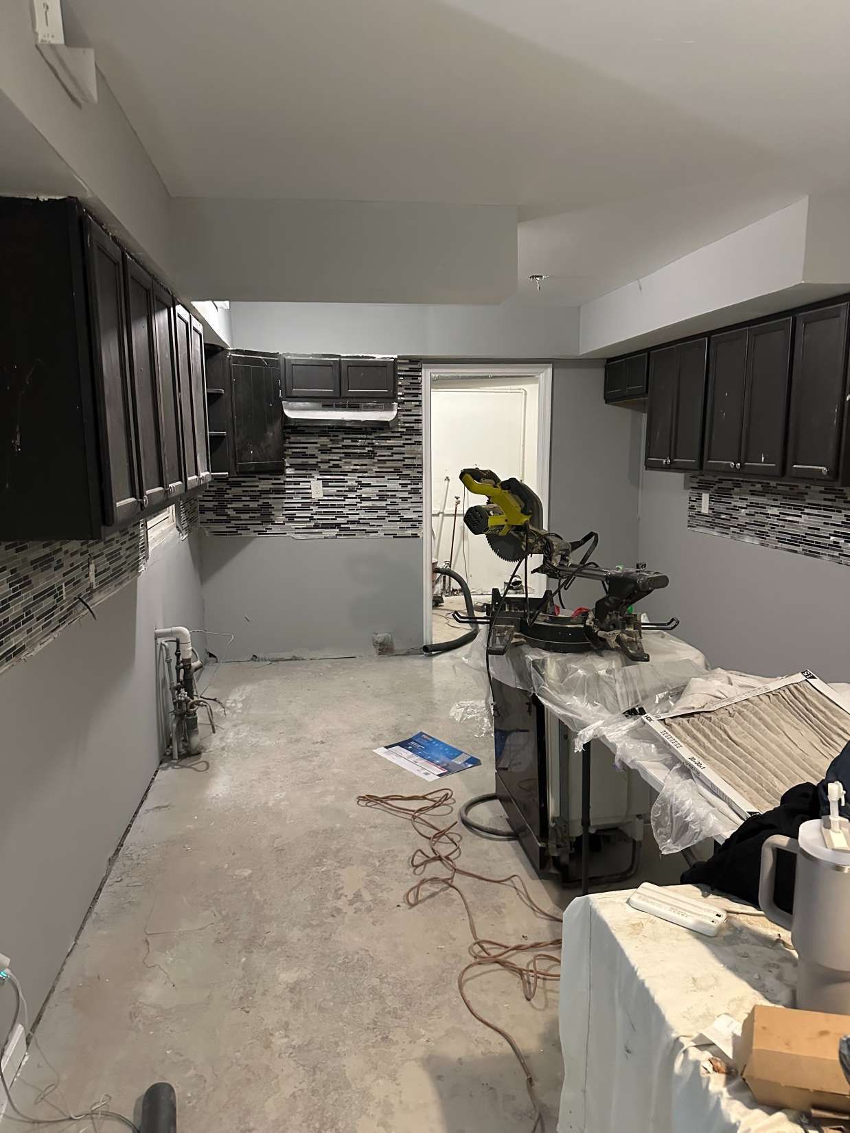 Narrow kitchen under renovation, with dark cabinets, tile backsplash, tools, and exposed floor wiring.