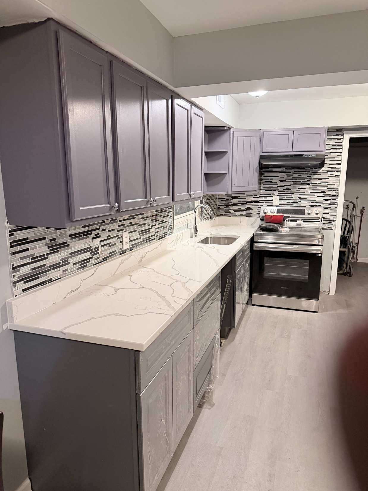 Small kitchen with gray cabinets, white countertops, patterned backsplash, and a stove at the far end.