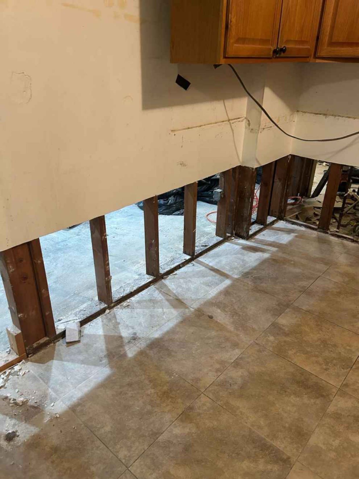 Damaged interior wall with exposed studs and wiring beneath a cabinet over a concrete floor