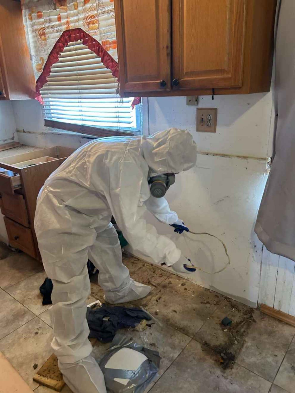 Person in white protective suit cleaning a damaged kitchen wall and floor during renovation.