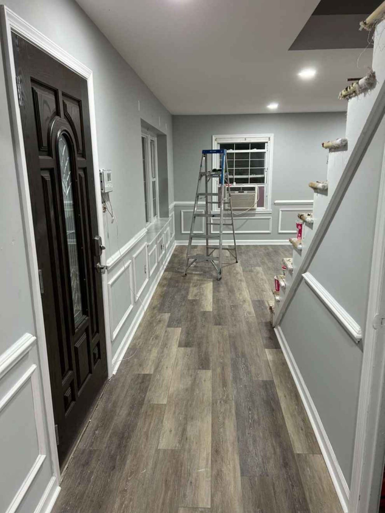 Narrow hallway with gray walls and wood-look flooring, featuring a folding ladder and window at the far end.