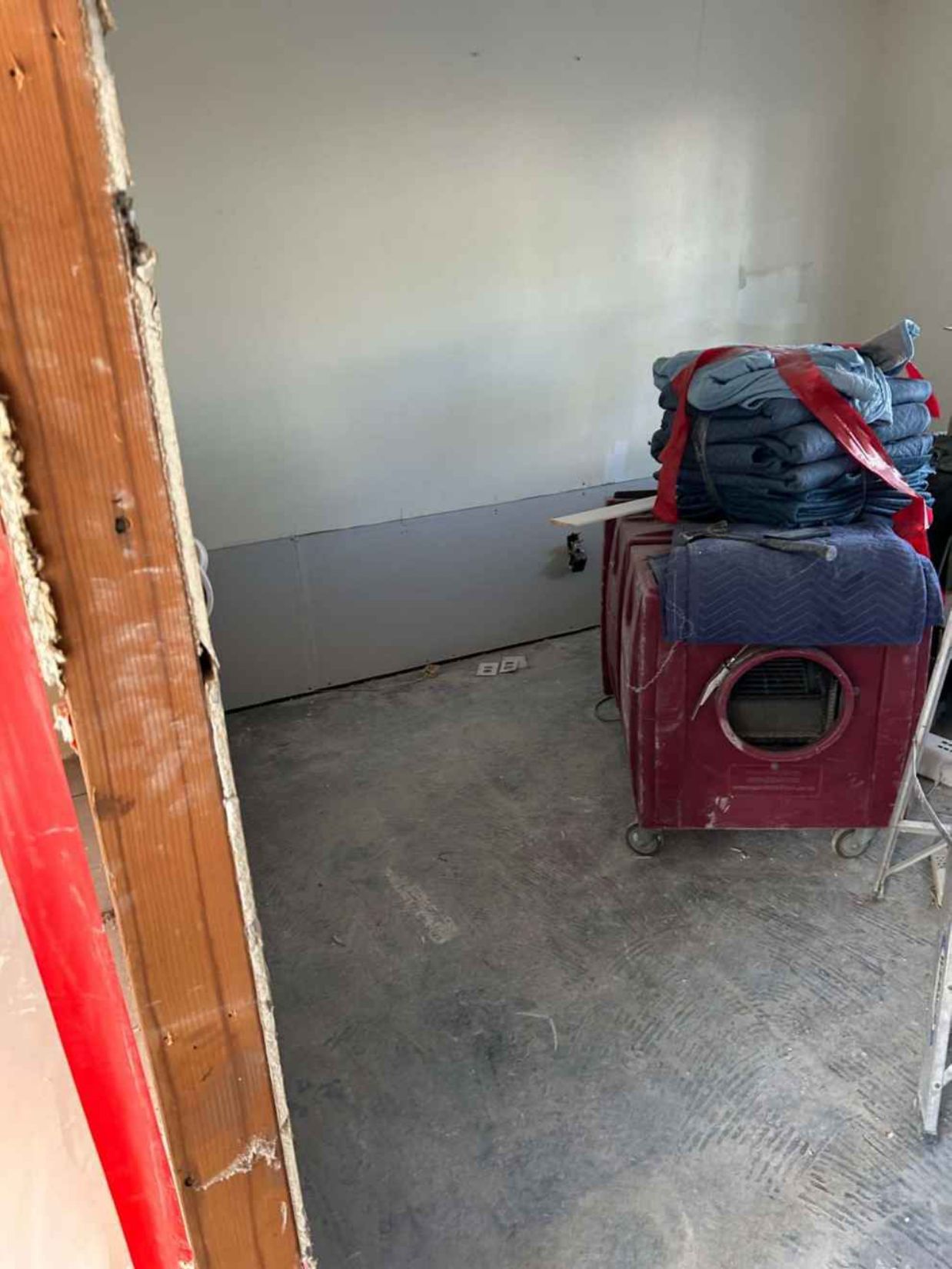 Small bare room with concrete floor, white walls, and stacked folded items on a red ottoman.