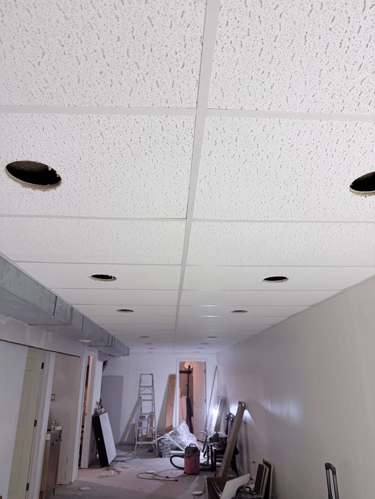 Narrow unfinished hallway with dropped ceiling, exposed lights, ladders, tools, and construction materials.