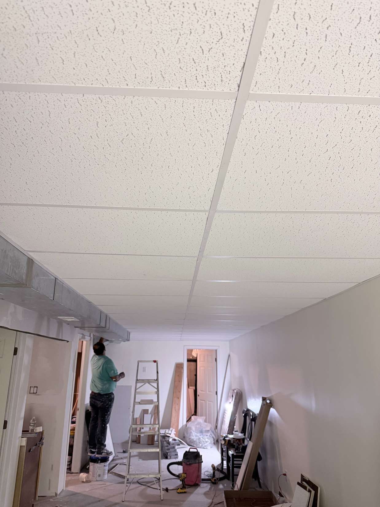 Worker on a ladder installing a drop ceiling in a narrow hallway under renovation.