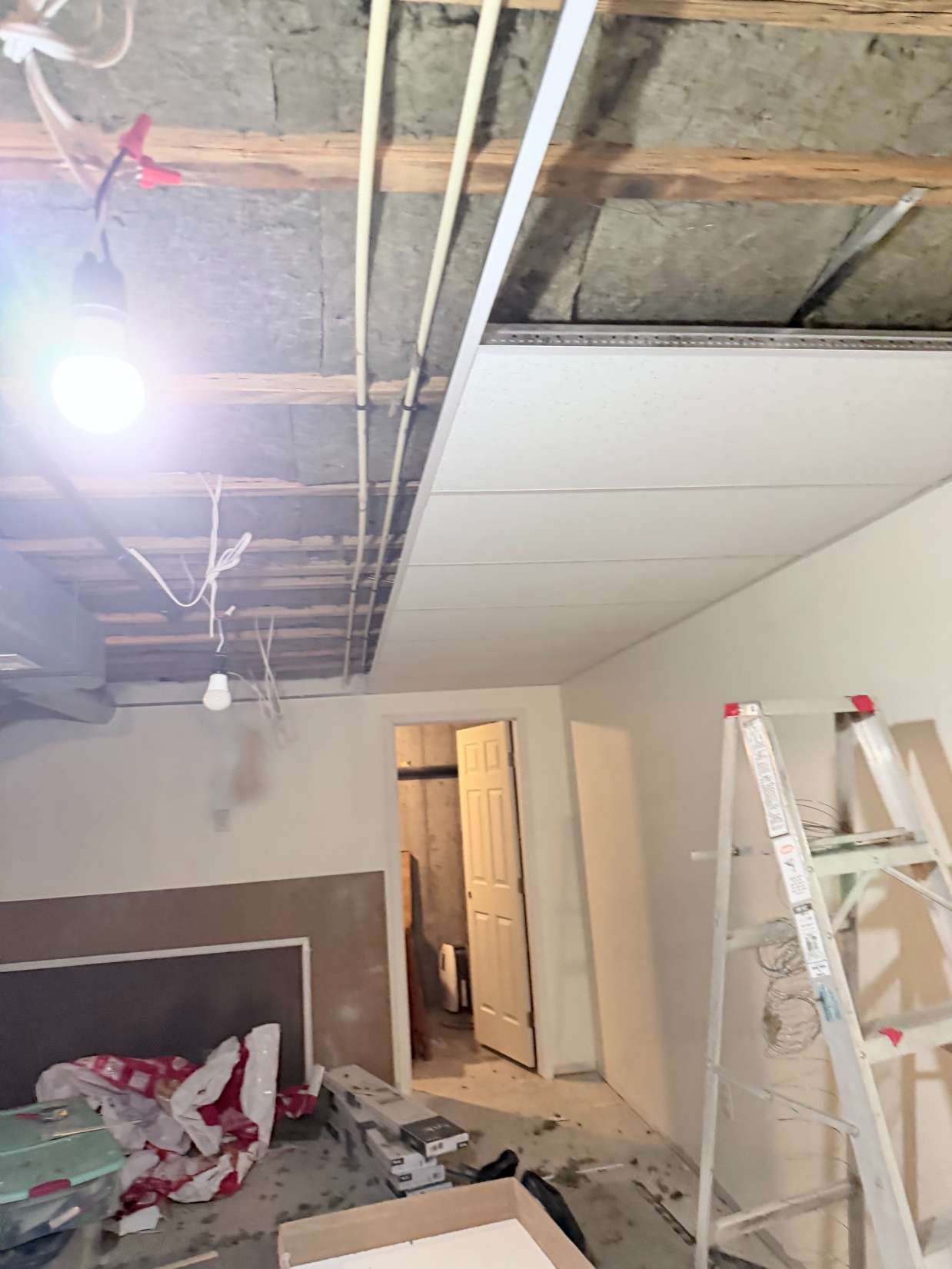 Partially renovated room with exposed ceiling, ladder, hanging lights, and debris on the floor