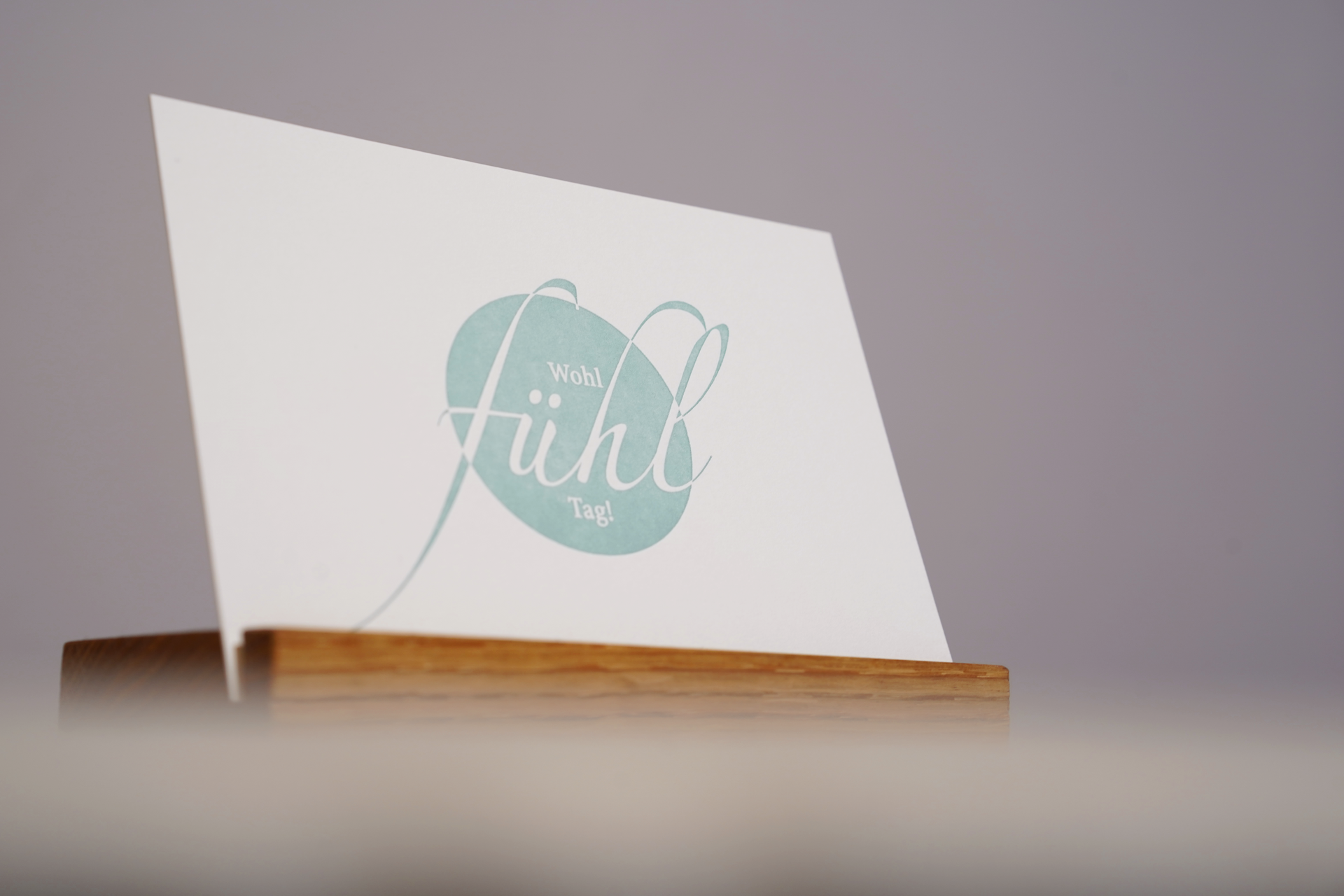 Weiße Tischkarte mit türkisfarbenem Blumenmonogramm auf einem Holztisch vor grauem Hintergrund