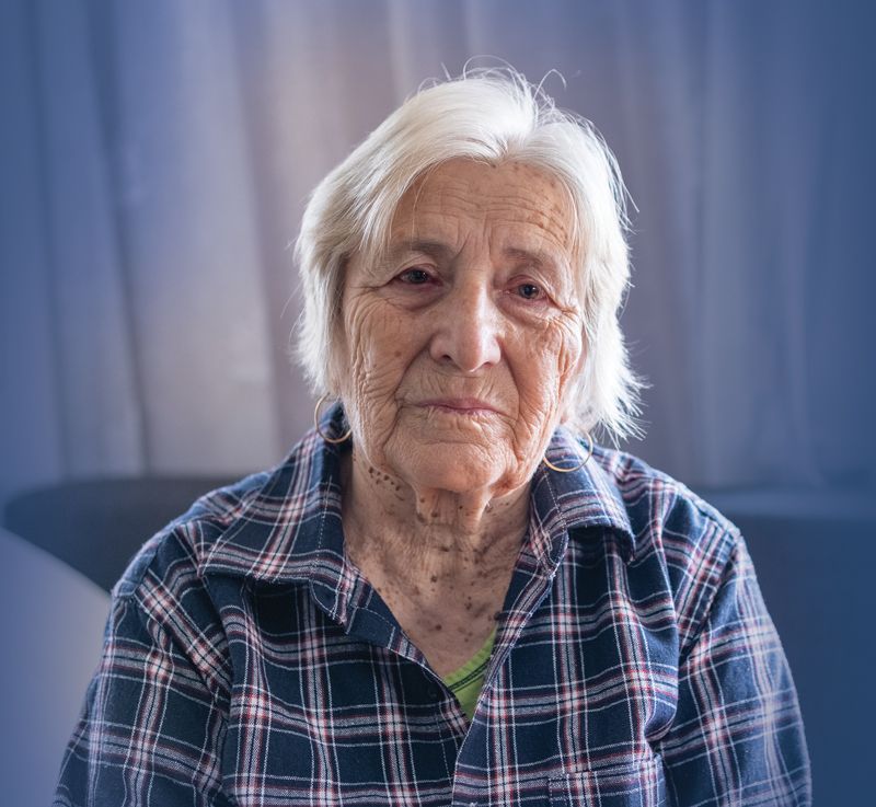 An elderly woman in a plaid shirt is sitting on a couch and looking at the camera.