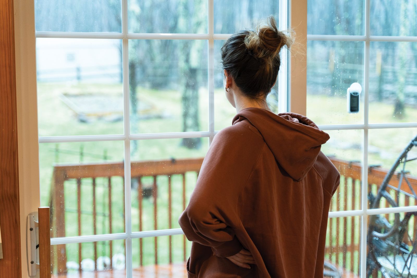 A woman in a brown hoodie is standing in front of a window looking out.