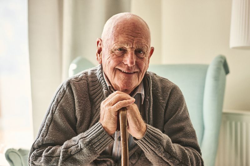 An elderly man is sitting in a chair with a cane and smiling.