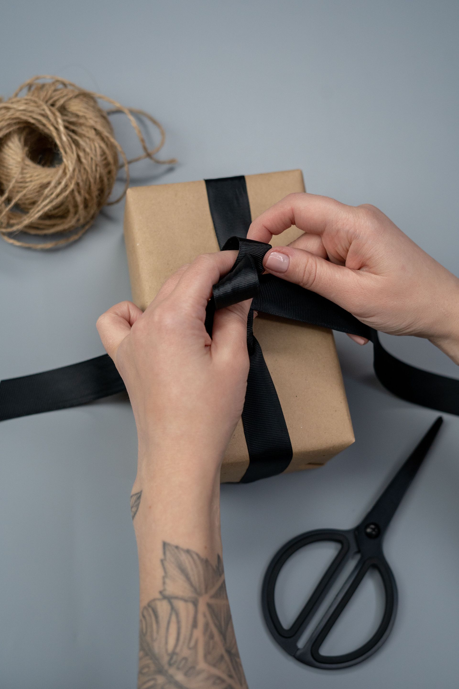 Handen die een zwart lint om een ​​klein ingepakt cadeautje knopen, naast een schaar en touw op een grijze achtergrond.