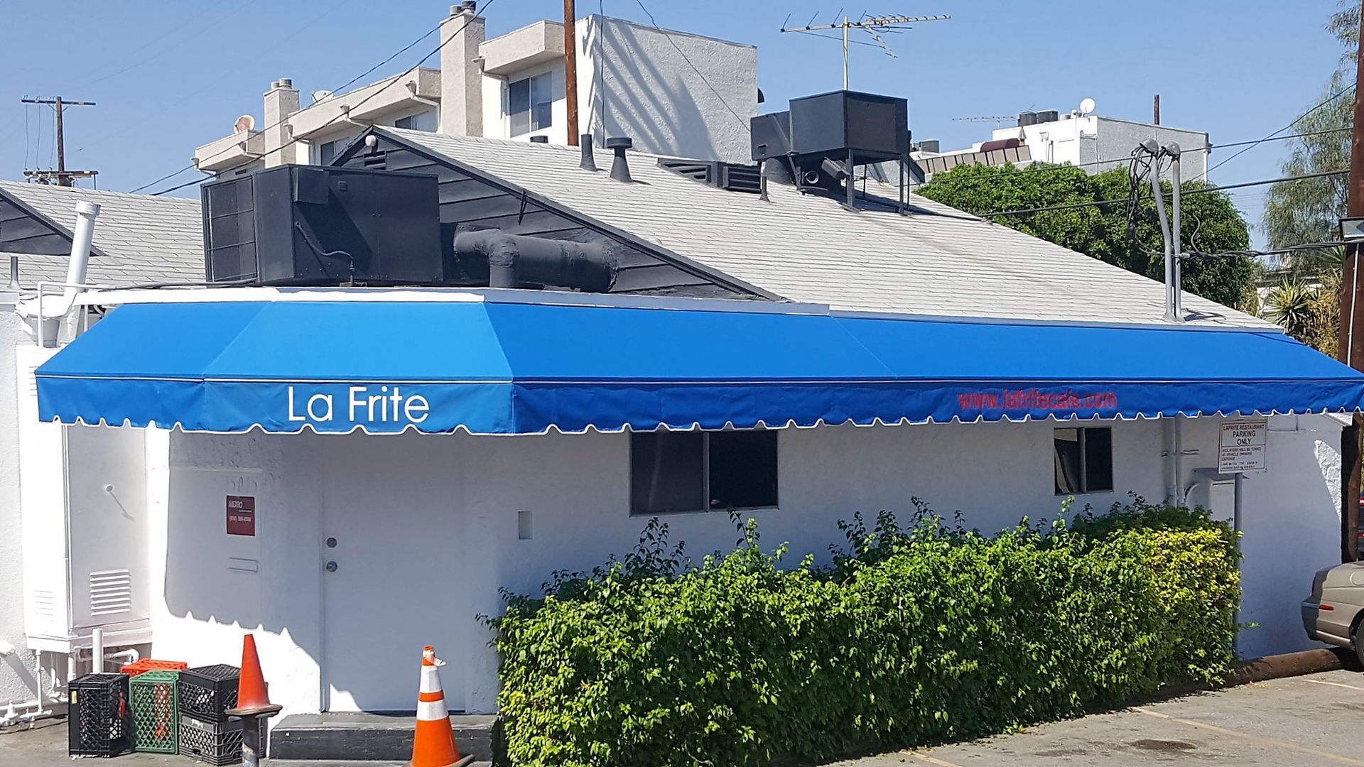 Blue awning over a small white storefront with hedge and traffic cones on a street corner