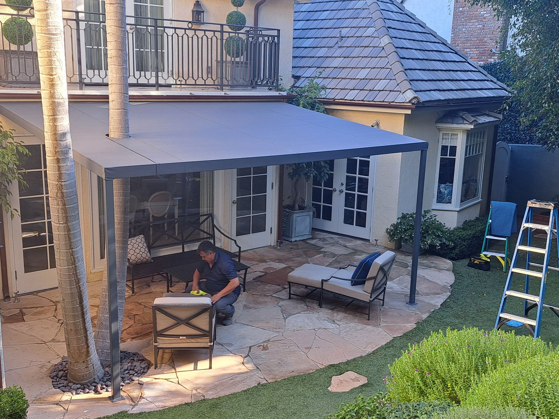 Patio with gray canopy, chairs, ladder, and a small white house with French doors.