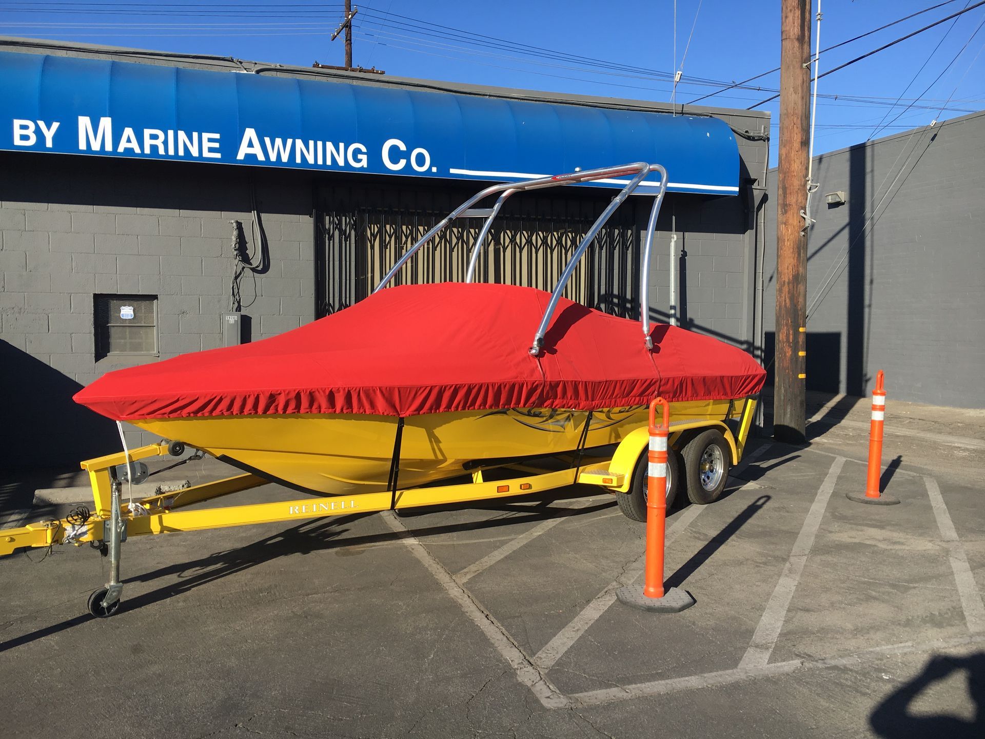 Red-covered boat on yellow trailer outside Marine Awning Co. building, with traffic cones nearby