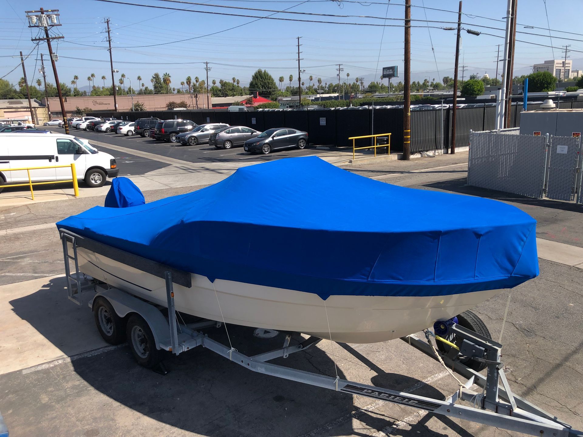 Blue-covered boat on a trailer in a parking lot, with cars and fences in the background.