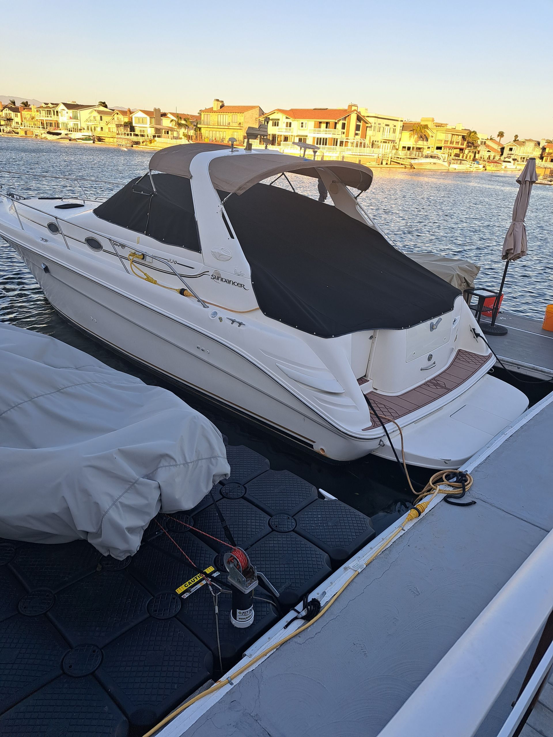 White yacht with black canopy docked beside a covered boat at a marina during sunset.