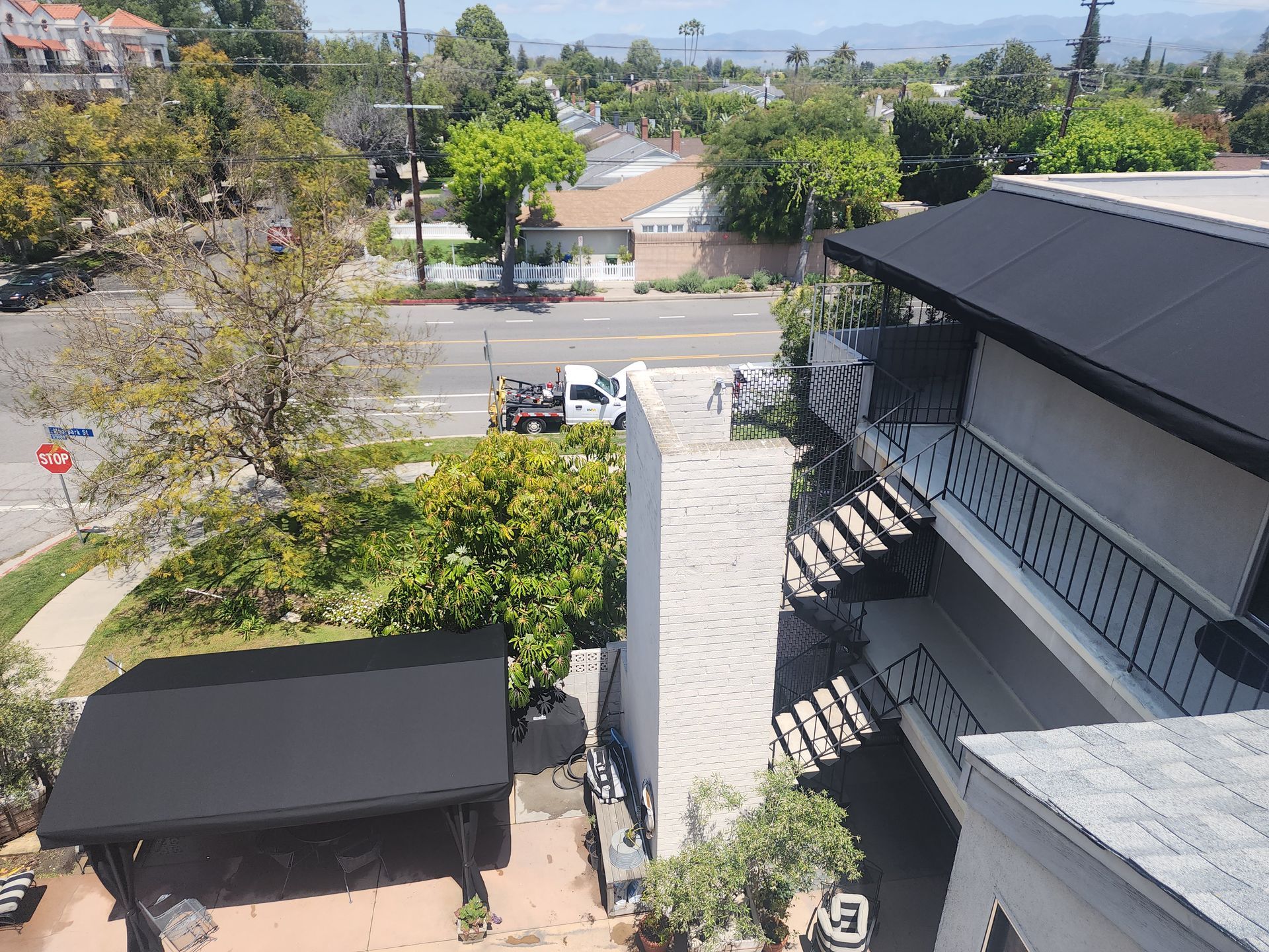 Residential street view from a balcony, with a stairway, trees, parked van, and rooftops on a sunny day