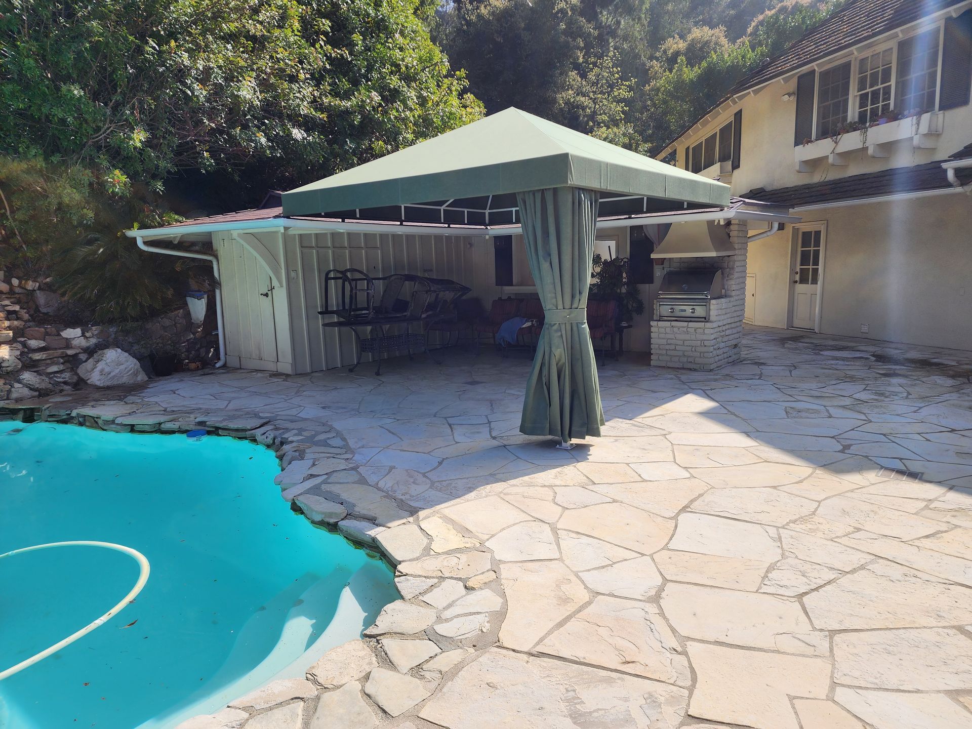 Poolside patio with a green canopy gazebo, stone paving, and a blue swimming pool beside a house