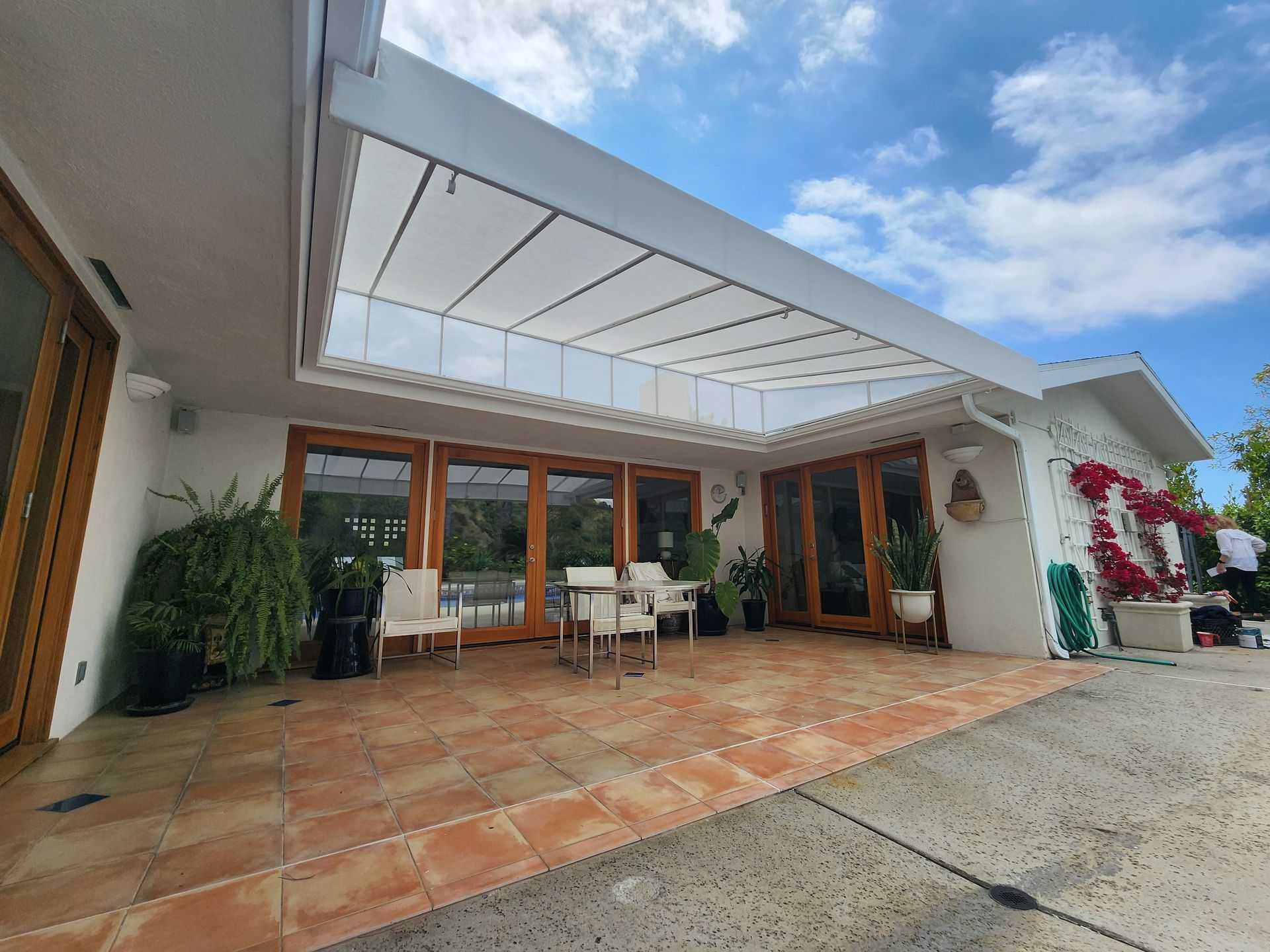 Covered patio with terracotta tiles, wooden doors, dining table, potted plants, and a blue sky overhead