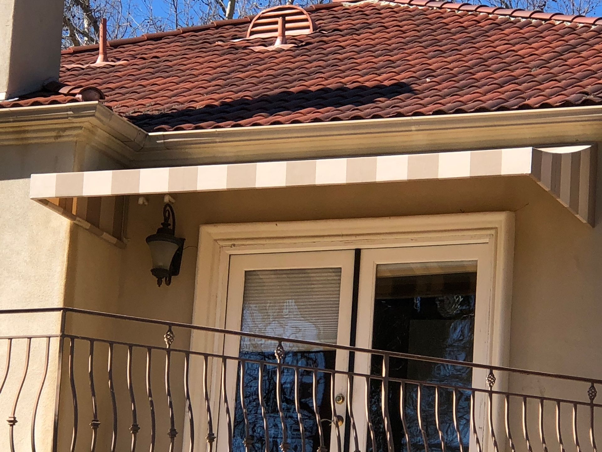Balcony with striped awning, cream door, black railing, and red-tiled roof on a house