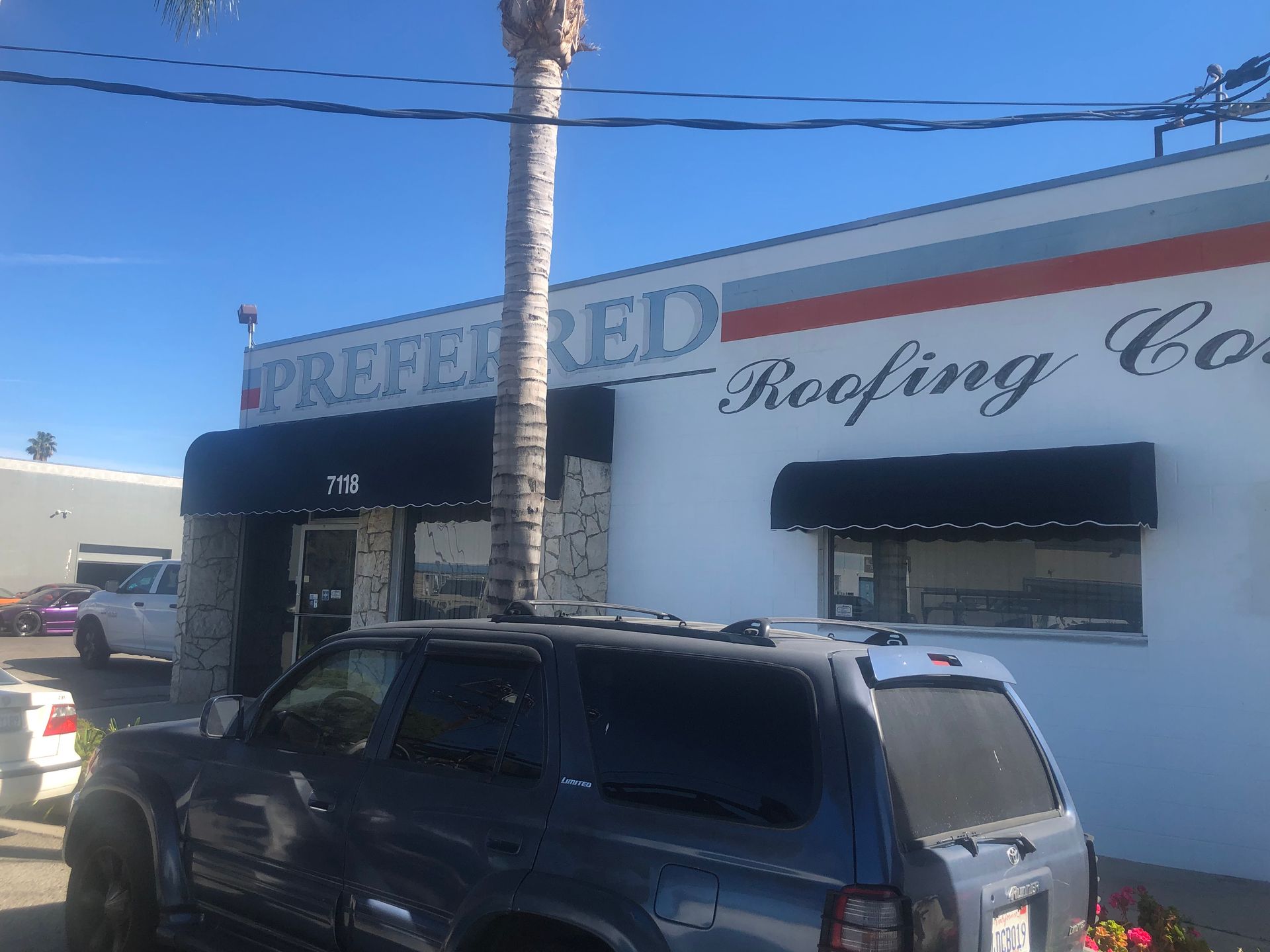 Roofing company building with parked SUV and blue awnings under a clear sky.
