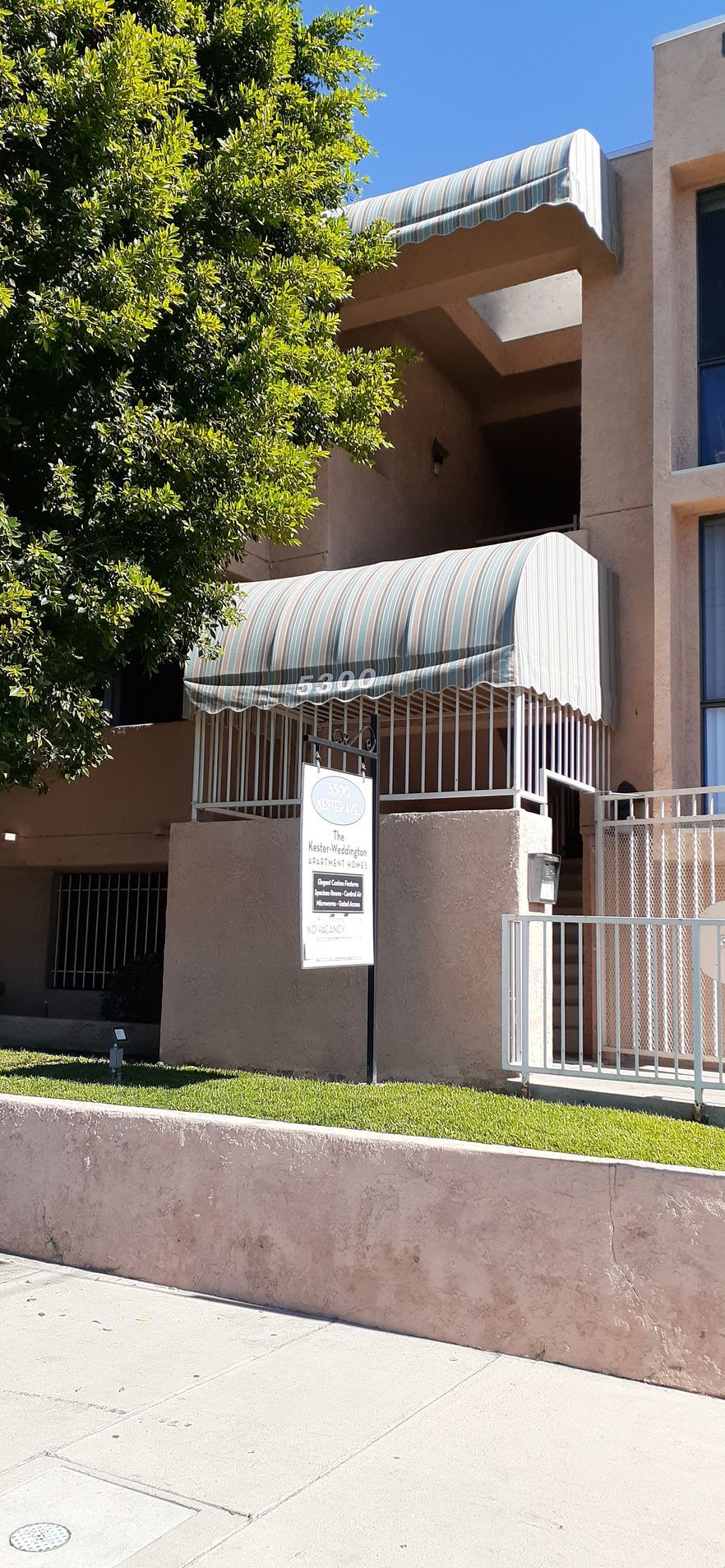 Apartment building entrance with white awning, railing, and large tree in front