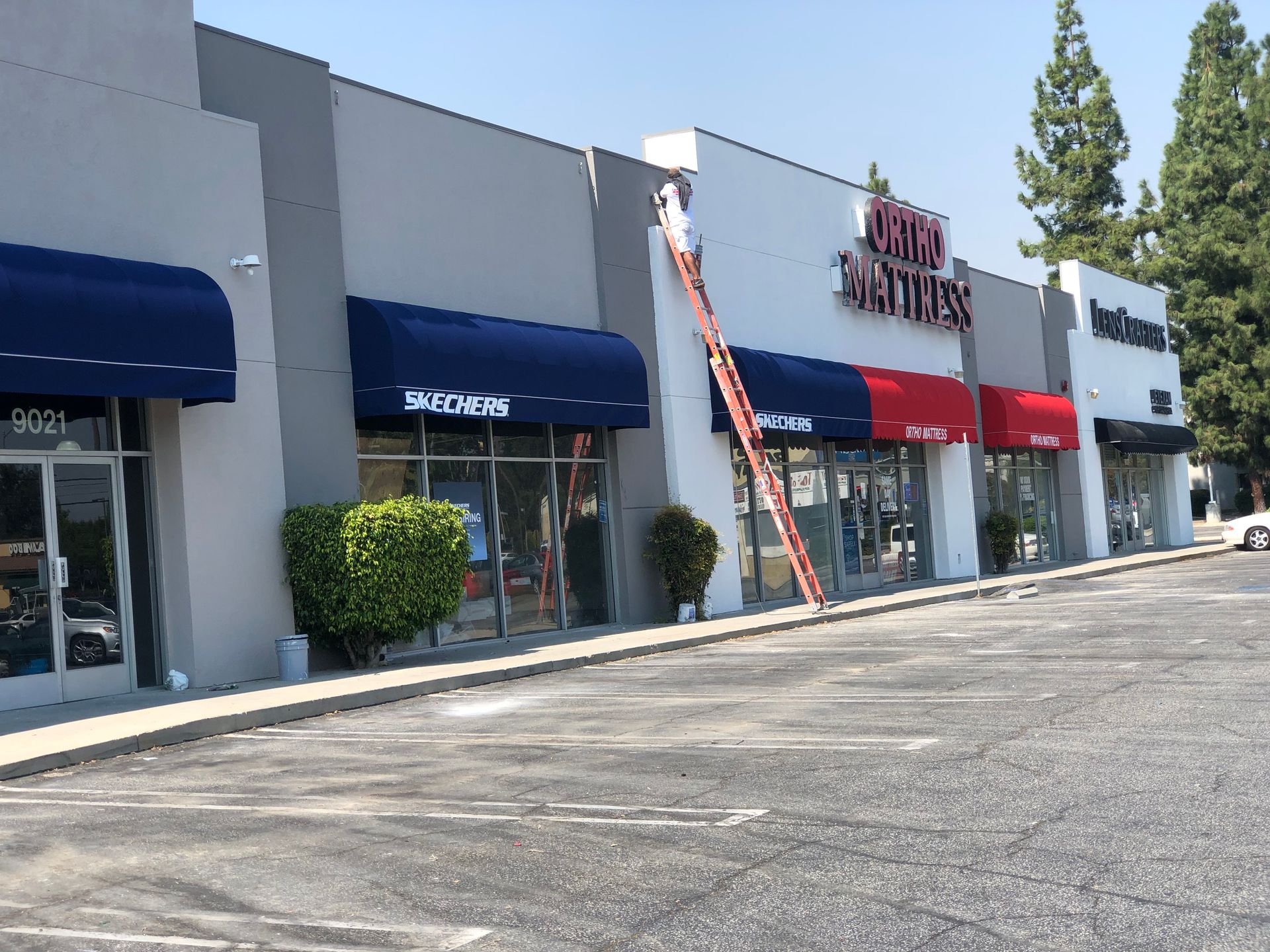 Shopping center storefronts with blue awnings and red signs, ladder leaning against the building, empty parking lot