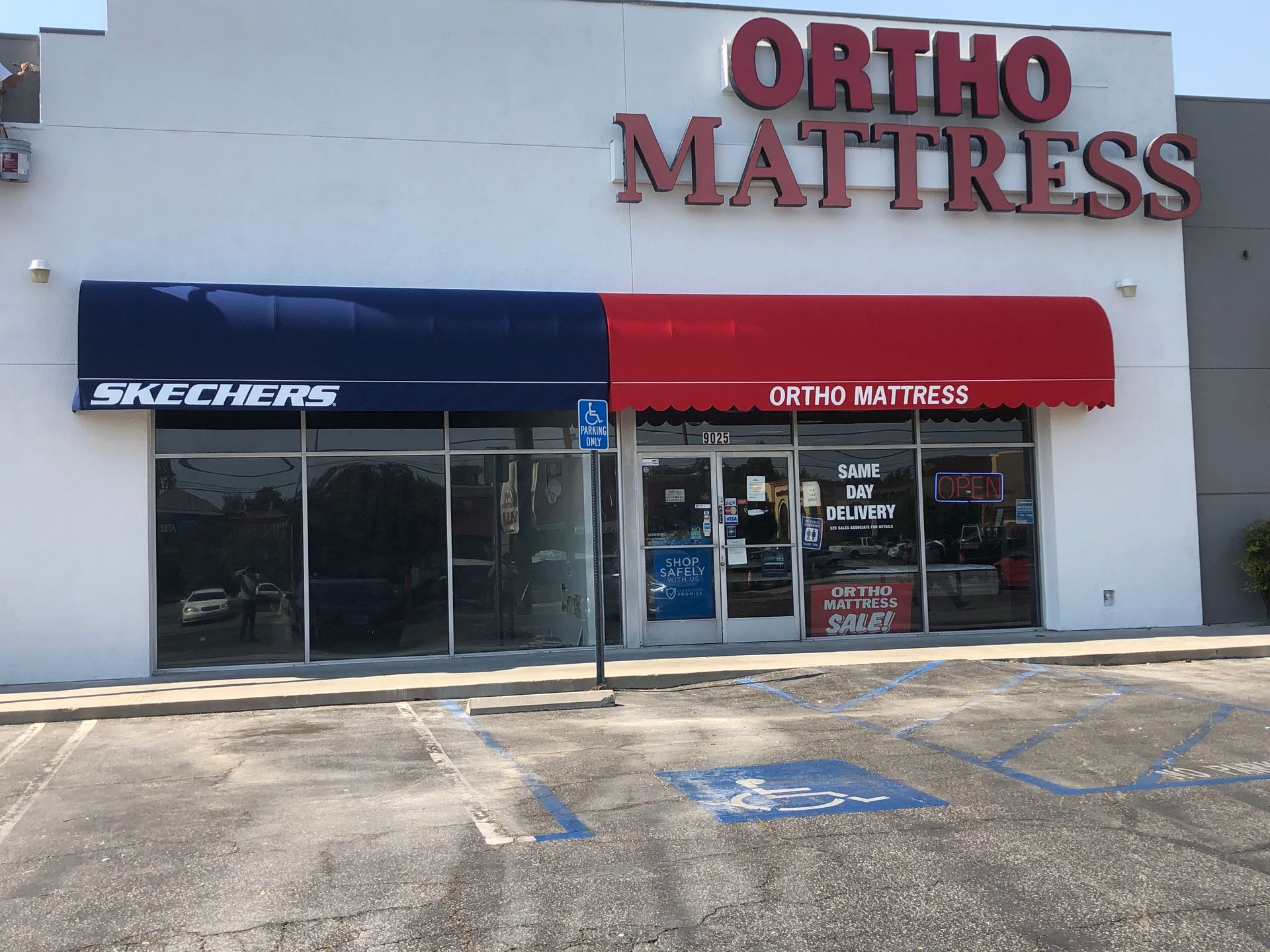 Storefronts of Ortho Mattress and Skechers in a strip mall, with empty parking spaces outside.